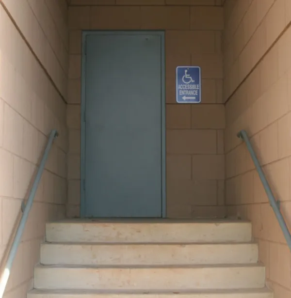 A sign with the universal symbol for access and an arrow points to a door with no handle at the top of stairs.