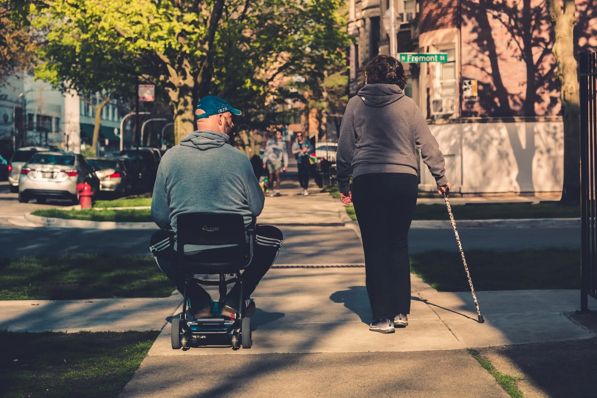 A man in a motorized chair and a woman with a cane walking down a sidewalk.